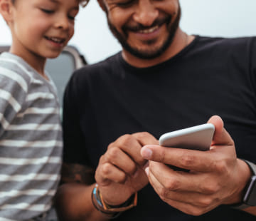 Imagem com homem utilizando celular e ao seu lado tem uma criança sorrindo